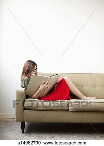 Stock Image - Young woman on sofa reading book. Fotosearch