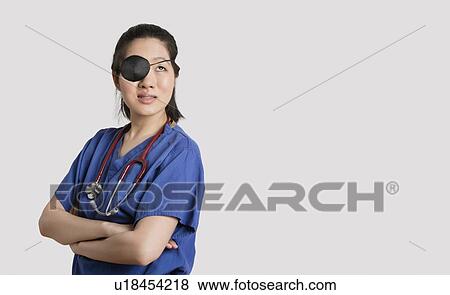 Stock Photo - Asian female doctor wearing an eye patch looking up with arms crossed over gray background. Fotosearch