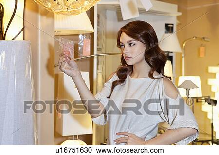 Stock Image - Beautiful woman looking at price tag of lighting fixture with hand on hip while standing in lights store. Fotosearch