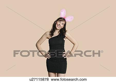 Beautiful young woman in black dress with bunny ears looking away over colored background View Large Photo Image Stock Photo - Beautiful young woman in black dress with bunny ears looking away over colored background. Fotosearch