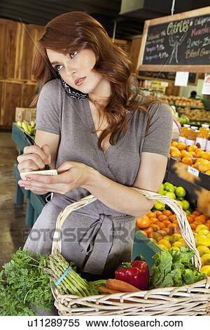 Stock Photography - Beautiful young woman listening to mobile phone while making a note of shopping list in market. Fotosearch