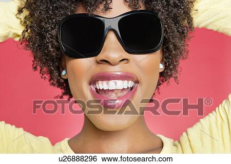 Stock Photograph - Close-up of a cheerful African American woman wearing sunglasses over colored background. Fotosearch