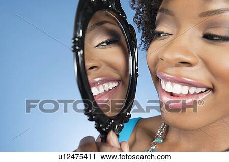 Close-up of a young woman looking at herself in mirror and smiling over colored background View Large Photo Image Stock Image - Close-up of a young woman looking at herself in mirror and smiling over colored background. Fotosearch