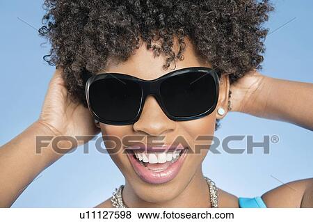 Stock Photo - Close-up of cheerful young woman wearing sunglasses with hands behind head over colored background. Fotosearch