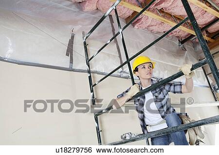 Stock Photograph - Female contractor climbing scaffold while looking away at construction site. Fotosearch