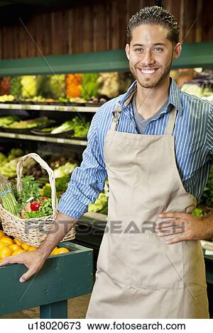 Stock Image - Handsome young sales clerk standing near stall in supermarket. Fotosearch