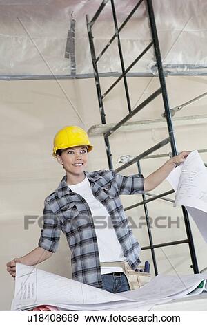 Happy mid adult worker holding building plans while looking away View Large Photo Image Stock Photo - Happy mid adult worker holding building plans while looking away. Fotosearch