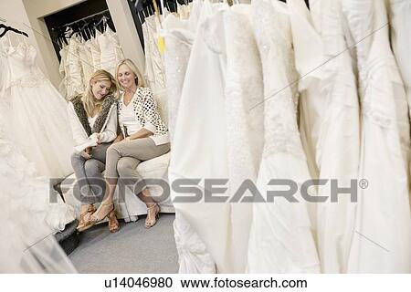 Stock Image - Happy mother and daughter sitting together on sofa in bridal boutique. Fotosearch