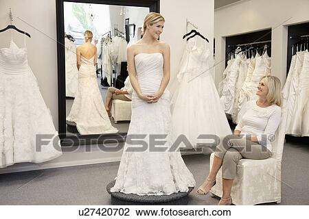 Stock Image - Happy mother looking at young daughter dressed in wedding gown in bridal boutique. Fotosearch