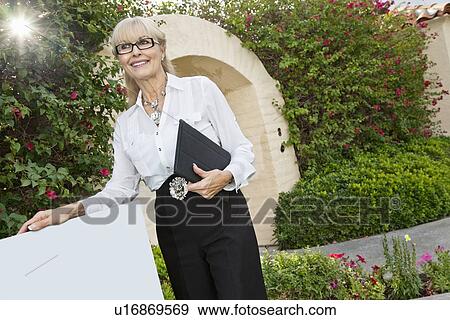Stock Photo - Happy senior woman standing by sign board with clipboard. Fotosearch