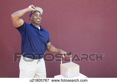 Stock Photo - Happy young African American delivery man standing with handtruck over colored background. Fotosearch