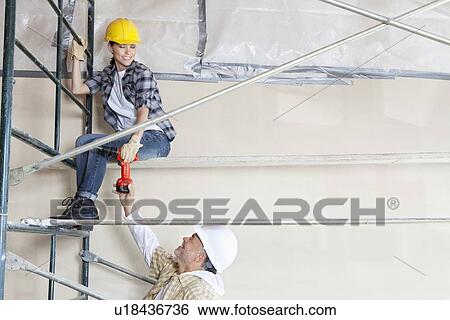 Stock Photograph - Male architect giving drill to female worker on scaffold at construction site. Fotosearch