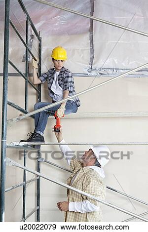 Stock Photo - Male worker giving drill to woman on scaffold at construction site. Fotosearch