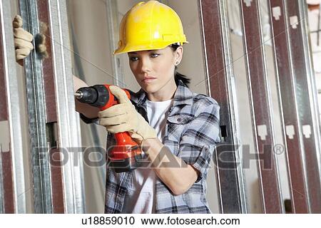 Stock Image - Mid adult female contractor drilling at construction site. Fotosearch