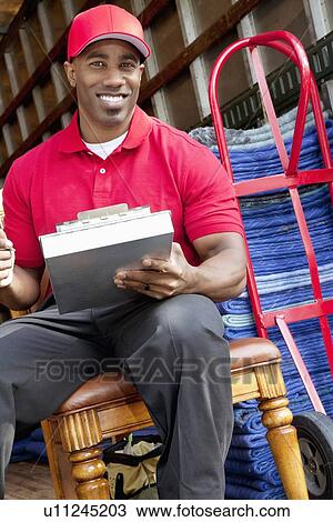 Stock Image - Portrait of a African American delivery man sitting on chair with clipboard. Fotosearch