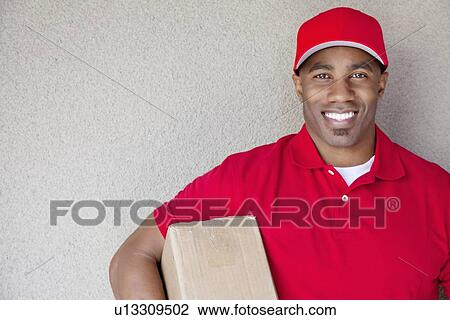 Stock Image - Portrait of a African American delivery man holding package against wall. Fotosearch