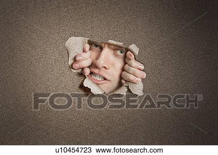 Stock Image - Portrait of a aggressive mid adult man peeking from ripped white paper hole. Fotosearch