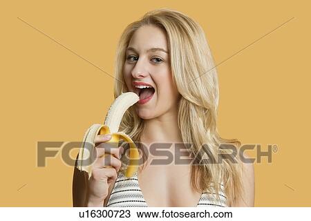 Stock Image - Portrait of a beautiful blond woman eating banana over colored background. Fotosearch