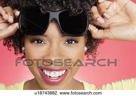 Stock Image - Portrait of a cheerful African American woman holding sunglasses over colored background. Fotosearch