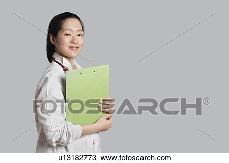 Stock Image - Portrait of a confident Asian female doctor holding clipboard over gray background. Fotosearch
