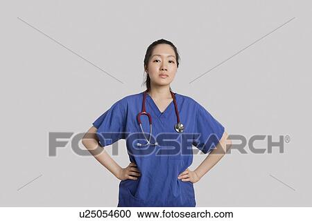 Stock Image - Portrait of a confident young Asian female doctor standing with hands on hips over gray background. Fotosearch