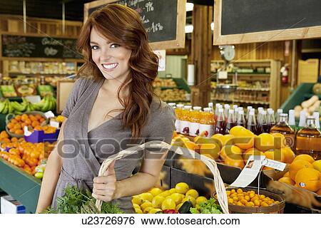 Stock Photograph - Portrait of a happy brunette woman standing with basket in fruit market. Fotosearch