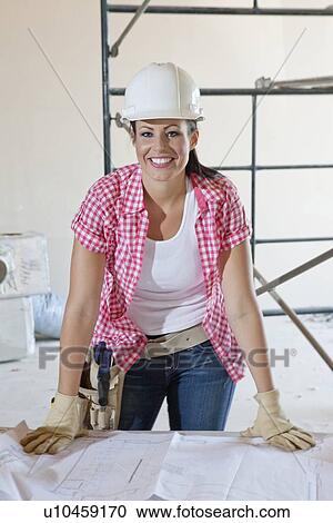 Stock Image - Portrait of a happy female construction worker wearing hardhat with blueprints. Fotosearch