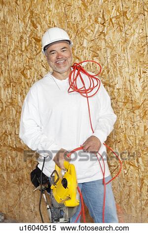 Stock Photography - Portrait of a happy male construction worker holding a power saw and a red electric wire. Fotosearch