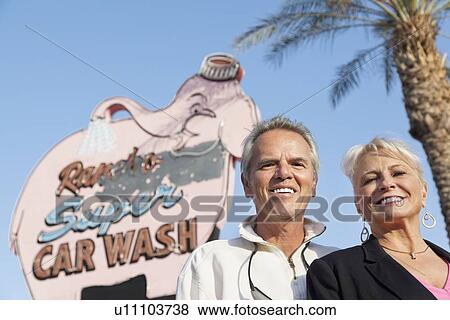 Stock Photo - Portrait of a happy mature couple in front of car wash signboard. Fotosearch