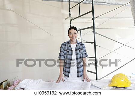 Stock Image - Portrait of a happy mid adult woman with building plans at construction site. Fotosearch