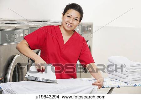 Picture - Portrait of a happy woman employee in red uniform ironing clothes in Laundromat. Fotosearch