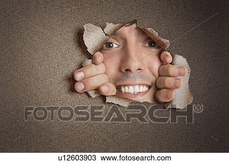 Stock Image - Portrait of a happy young Hispanic man peeking from ripped paper hole. Fotosearch
