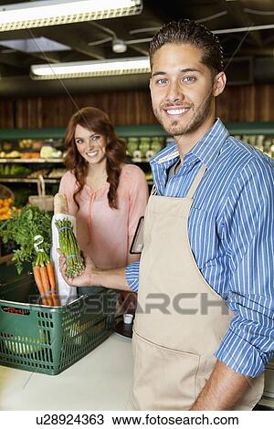 Stock Image - Portrait of a happy young sales clerk holding vegetables with woman in background. Fotosearch