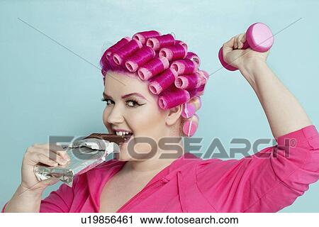 Portrait of a happy young woman biting chocolate bar while lifting dumbbell over colored background View Large Photo Image Stock Image - Portrait of a happy young woman biting chocolate bar while lifting dumbbell over colored background. Fotosearch