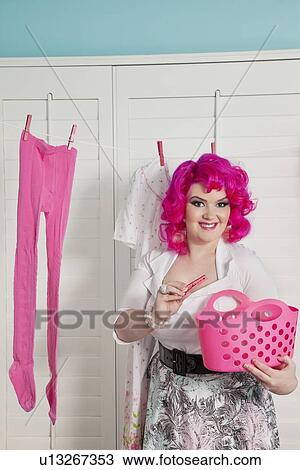 Stock Image - Portrait of a happy young woman with clothespin hanging clothes in room. Fotosearch