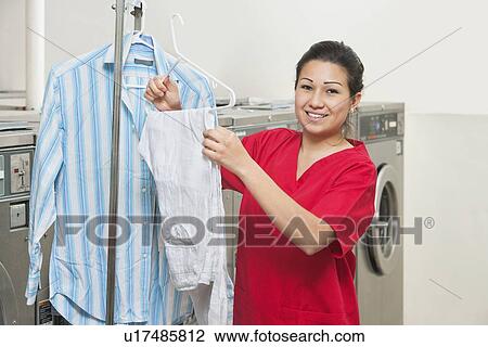 Stock Image - Portrait of a happy young woman with shirt hanging in Laundromat. Fotosearch