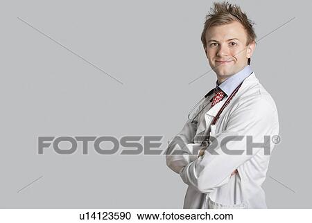 Stock Image - Portrait of a male doctor smiling with arms crossed over gray background. Fotosearch