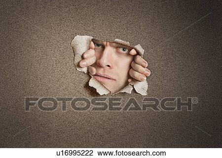 Stock Image - Portrait of a mid adult Caucasian man peeking from ripped white paper hole. Fotosearch