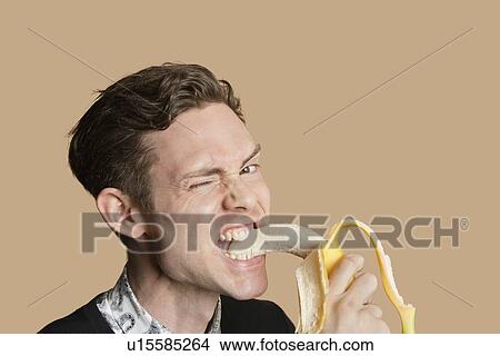Picture - Portrait of a mid adult man winking while biting banana over colored background. Fotosearch