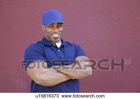 Stock Image - Portrait of a muscular African American delivery man with arms crossed over colored background. Fotosearch