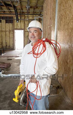Stock Photo - Portrait of a smiling male construction worker holding a power saw and a red electric wire. Fotosearch