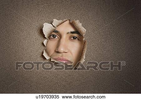 Stock Photography - Portrait of a young Indian man peeking from ripped paper hole. Fotosearch