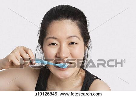 Stock Photography - Portrait of a young woman brushing her teeth over light gray background. Fotosearch