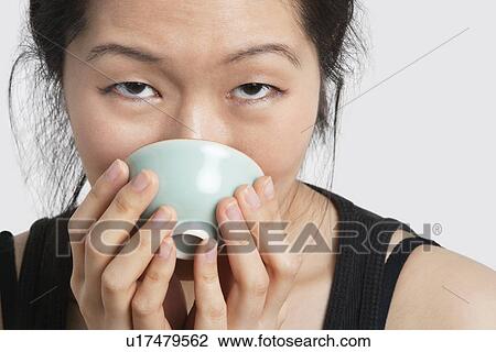 Stock Image - Portrait of a young woman drinking from bowl over light gray background. Fotosearch
