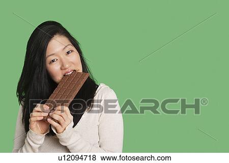 Stock Photo - Portrait of a young woman eating a large chocolate bar over green background. Fotosearch