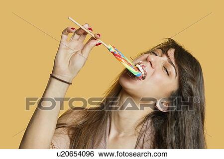 Stock Photograph - Portrait of a young woman eating lollipop over colored background. Fotosearch