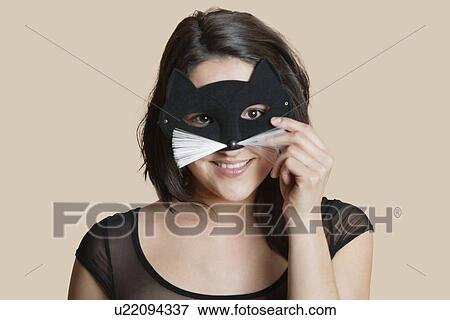 Stock Photo - Portrait of a young woman looking through eye mask over colored background. Fotosearch