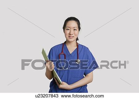 Stock Image - Portrait of an Asian female nurse holding clipboard over gray background. Fotosearch