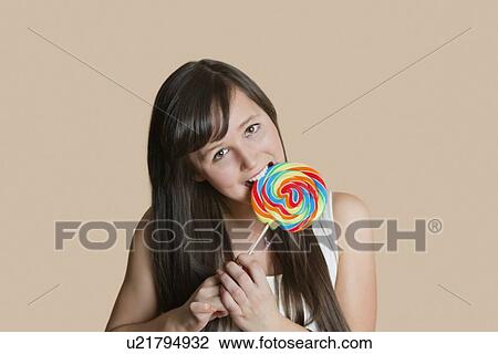 Stock Image - Portrait of beautiful young woman biting lollipop over colored background. Fotosearch
