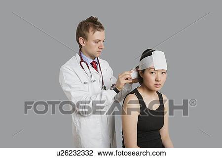 Stock Image - Portrait of female patient with doctor wrapping bandage to her head. Fotosearch